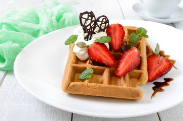 Whole wheat Belgian waffles with whipped cream, freshly chopped strawberries, mint leaves and chocolate on white wooden background. Homemade healthy breakfast.