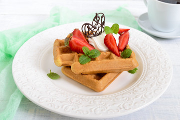 Whole wheat Belgian waffles with whipped cream, freshly chopped strawberries, mint leaves and chocolate and a cup of tea on white wooden background. Homemade healthy breakfast.