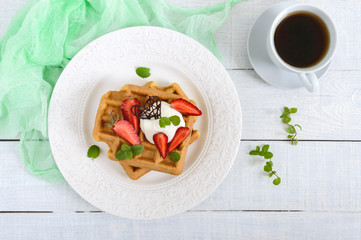 Whole wheat Belgian waffles with whipped cream, freshly chopped strawberries, mint leaves and chocolate and a cup of tea on white wooden background. Homemade healthy breakfast. The top view