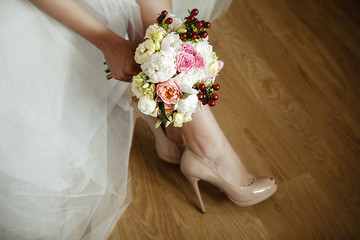 Closeup view of female legs in beige high heels and in the hands of a wedding bouquet of peonies. Beautiful young woman in long gorgeous dress indoors. Horizontal color photo.