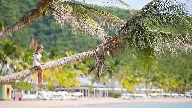 Adorable little girl sitting on palm tree during summer vacation on white beach