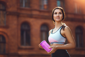 Young attractive sports woman drinking sport supplements after running, working out or exercising outdoors. Brunette holding shaker and looking away from the camera. Fitness girl during sport routine