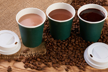 Three take away green paper cups with coffee beans and hot chocolate drink on wooden background, top view, close-up