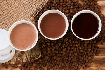 Three take away green paper cups with coffee beans and hot chocolate drink on wooden background, top view, close-up