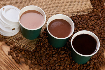 Three take away green paper cups with coffee beans and hot chocolate drink on wooden background, top view, close-up