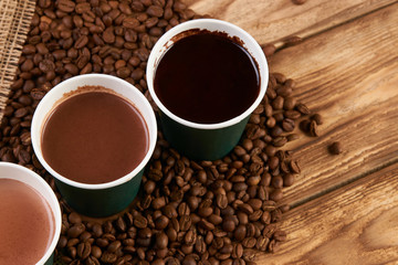 Three take away green paper cups with coffee beans and hot chocolate drink on wooden background, top view, close-up