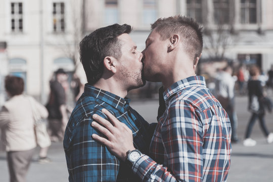 gay couple in the street of a city kissing each other passers by in the background. their are hugging each other passionate