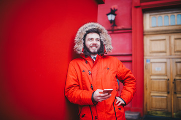 Fototapeta premium handsome young male student with toothy smile and beard stands on red wall background in bright red winter jacket with hood with fur, Uses, holds mobile phone with hand in pocket. Winter cold weather
