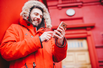 handsome young male student with toothy smile and beard stands on red wall background, facade of educational institution in red winter jacket with hood with fur, Uses finger on screen of mobile phone