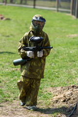 Boy in the camouflage holds a paintball gun  in one hand and protective helmet , standing on the field with group of players on the background