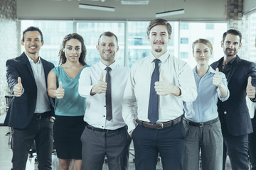 Group of successful young business people standing in office, showing thumbs up sign and smiling. Business success concept