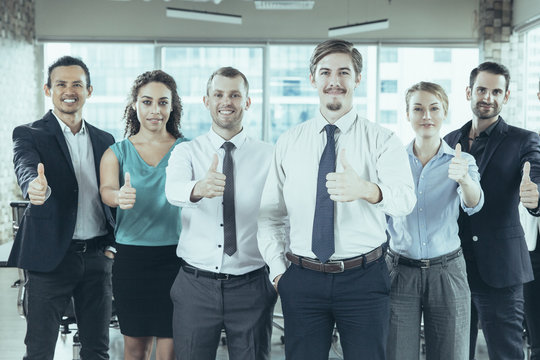 Group of successful young business people standing in office, showing thumbs up sign and smiling. Business success concept
