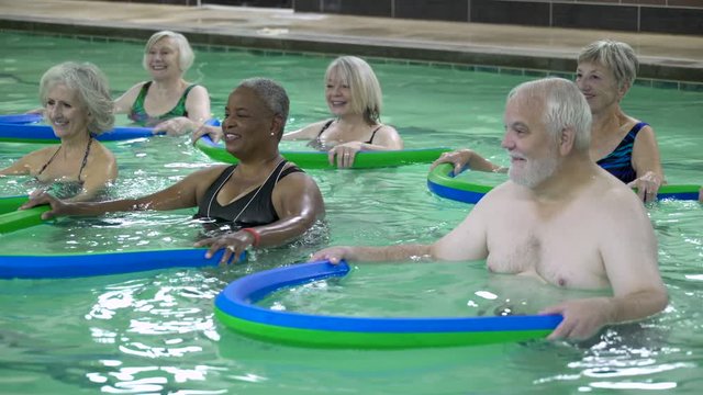 Mature adults exercising with pool noodles in swimming pool.