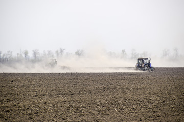 Fototapeta premium The tractor harrows the soil on the field and creates a cloud of dust behind it