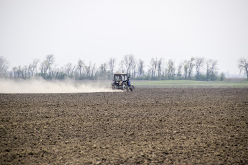 The tractor harrows the soil on the field and creates a cloud of dust behind it