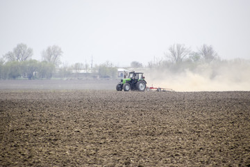 The tractor harrows the soil on the field and creates a cloud of dust behind it