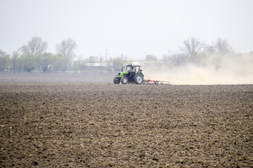The tractor harrows the soil on the field and creates a cloud of dust behind it