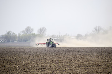 The tractor harrows the soil on the field and creates a cloud of dust behind it