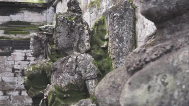 Sculpture near Holy Fauntain in the wild tropical jungle, detailing the weathered surface with white fungus and moss. Lush vegetation and Balinese temple on the background. Slow tracking shot