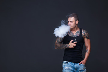 Vaper. The man with a muscular torso with tattoos smoke an electronic cigarette on the dark background