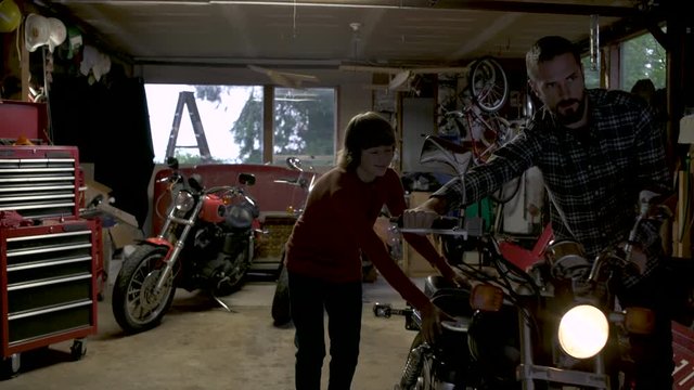 Father and teenage son pushing motorbike in garage. 