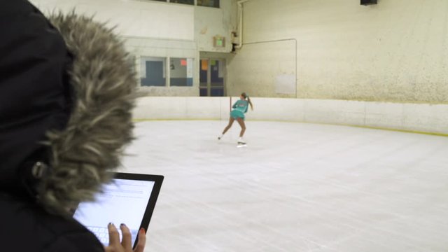Young girl ice skating on ice rink with woman using digital tablet.