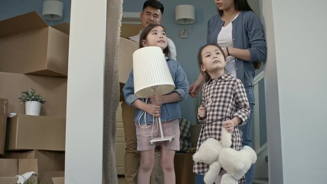 Tilt up of happy little girl with plush toy and her sister with lamp looking around their new house and talking with cheerful parents after moving
