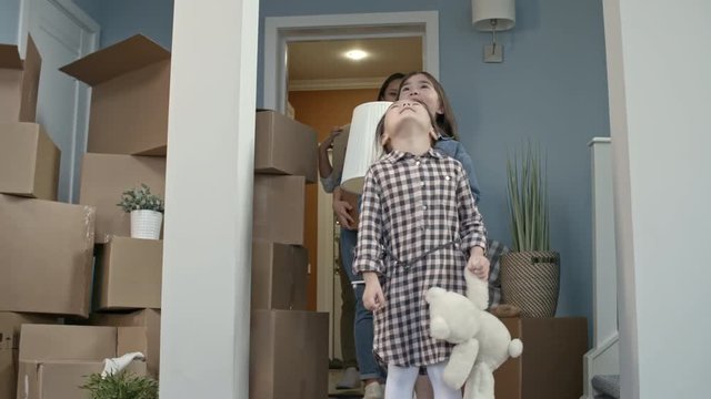 PAN of happy little girls with plush toy and lamp running into new house and looking around in amazement as their parents carrying cardboard box and carpet smiling