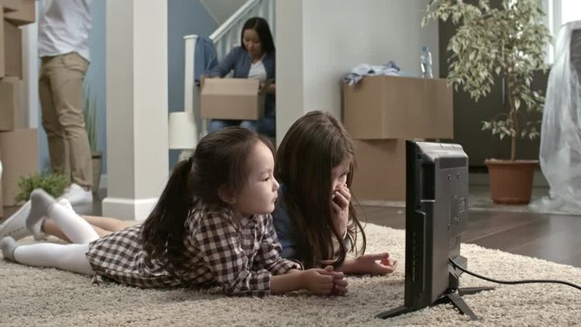 PAN of cute little girls lying on carpet empty living room of new house and watching TV as their parents unloading stuff and unpacking cardboard boxes in background