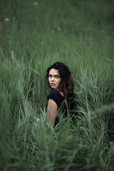 Naklejka na ściany i meble Pretty woman sitting on the grass in dark dress. Beautiful woman posing in tall grass. Brunette lady in field at sunset. Freedom concept background