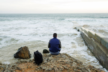 the guy is sitting with his back against the sea