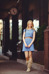 beautiful blond woman standing under the house with a large clock dressed in a blue dress and boots, modest and shy