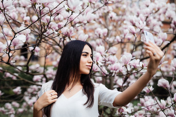 Beautiful stylish caucasian woman making selfie in blossom magnolia garden. Spring time