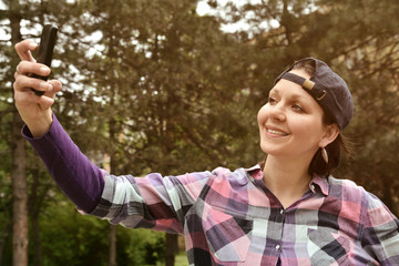 Beautiful woman wearing checked shirt and baseball cap taking selfie in the city park. Selective focus.