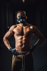 Fototapeta na wymiar african american athletic man in mask looking at camera and holds hands on belt. black man posing in gym. black background