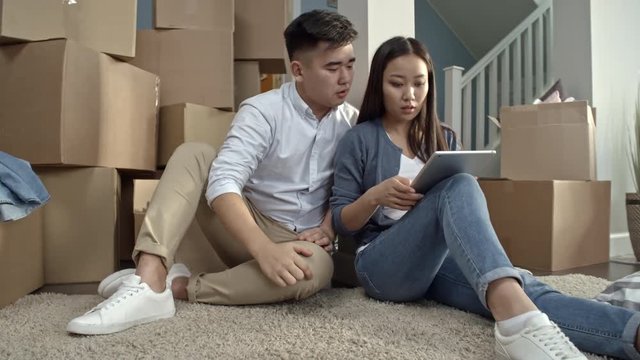 PAN of Asian couple sitting on floor of their new apartment and looking at tablet while choosing furniture layout