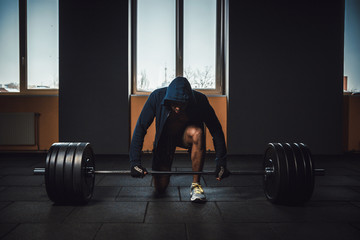 athletic man in jacket with a hood waiting and preparing before lifting heavy barbell. fitness, sport, training, gym and lifestyle concept. deadlift with barbell in mask. bent over the barbell