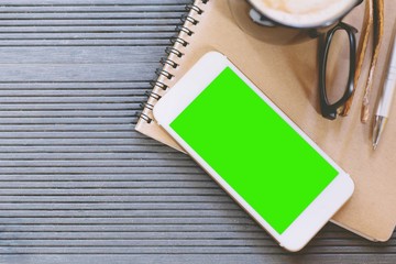 mobile smart phone display green screen on office desk table with book notepad,equipment supplies in the work and coffee cup. Top view with copy space. concept to work in a modern office.