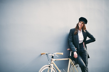 Woman posing in front of wall yellow bicycle behind her.. Hand holding hat.