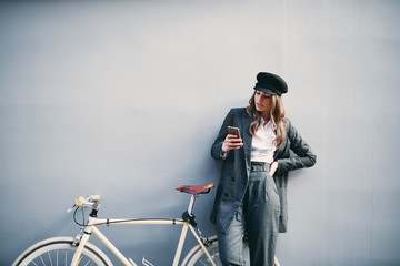 Woman standing against the wall and using smart phone for texting. Next to her yellow bicycle.
