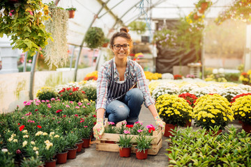 Beautiful smiling florist woman posing while crouching and holding the wooden box with flowerpots in the colourful bright greenhouse.