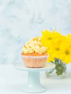 Cupcake with yellow cream decoration on stand and bouquet of chrysanthemum in glass vase on blue pastel background.