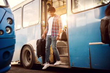 Young joyful man is heading down the bus stairs while carrying a traveling bag.