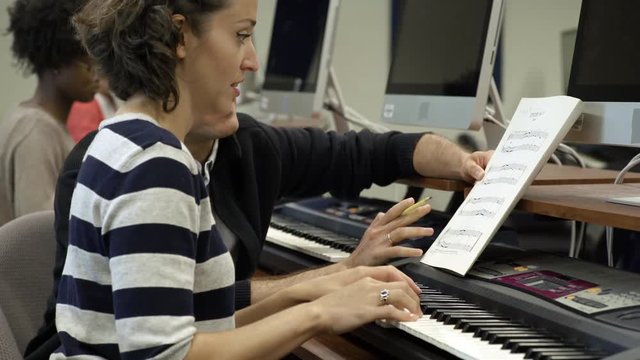 Woman learning to play electric keyboard in college music lesson with tutor helping.