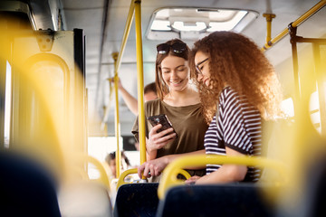 Two cheerful pretty young women are standing in a bus and looking at the phone and smiling while...