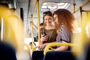 Foto op Canvas Two cheerful pretty young women are standing in a bus and looking at the phone and smiling while waiting for a bus to take them to their destination. © dusanpetkovic1