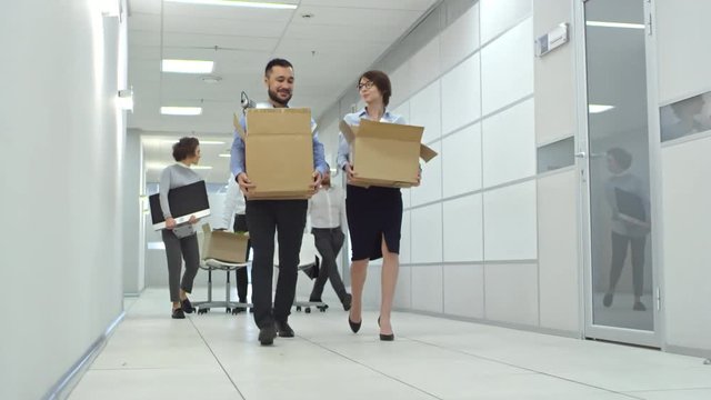 Group of cheerful office workers walking through hallway towards the camera while moving into new office. Colleagues carrying cardboard boxes with belongings, computer and chairs