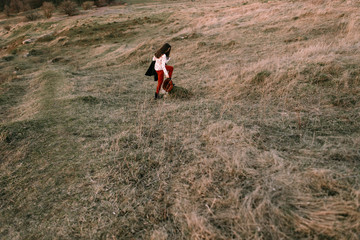 Stylish traveler woman with hat holding map on top of mountains, emotional happy moment, travel concept, space for text. Girl  walking along the grass at the top of the mountain in sunset