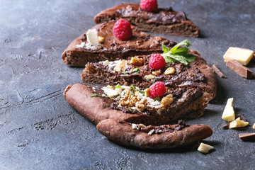 Sliced homemade sweet dessert chocolate pizza with different chocolates, raspberries and mint served with ingredients above over blue texture background.