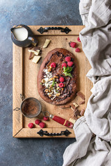 Homemade sweet dessert chocolate pizza with different chocolates, raspberries and mint served on wooden tray with cloth and ingredients above over blue texture background. Top view, space.